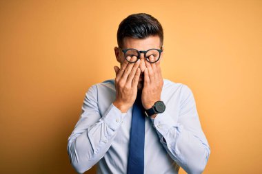 Sarı arka planda duran kravat ve gözlük takan genç yakışıklı iş adamı yorgunluk ve baş ağrısı için gözlerini ovuşturuyor, uykulu ve yorgun ifade ediyor. Görüş sorunu