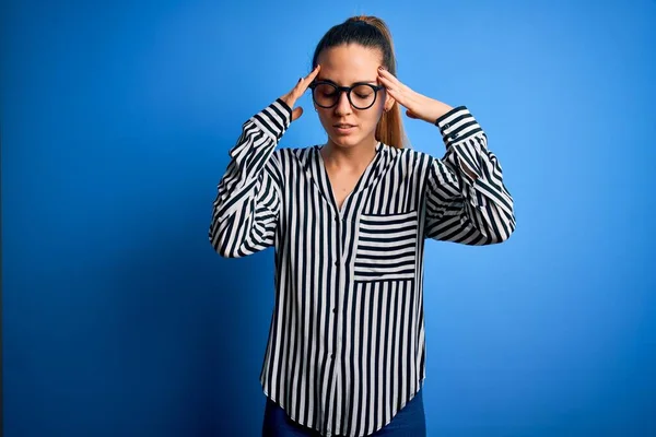 Beautiful blonde woman with blue eyes wearing striped shirt and glasses over blue background with hand on head for pain in head because stress. Suffering migraine.