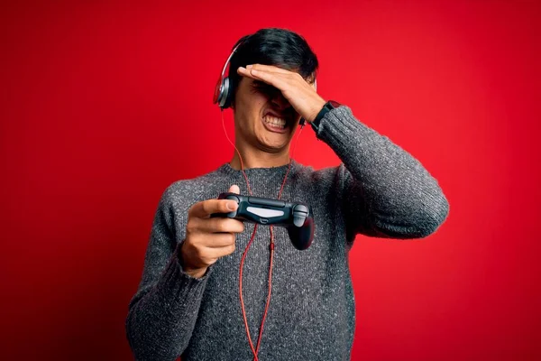 Young handsome gamer man playing video game using joystick and headphones stressed with hand on head, shocked with shame and surprise face, angry and frustrated. Fear and upset for mistake.