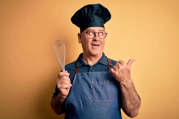 Middle age hoary cooker man wearing apron and hat holding whisk over yellow background pointing and showing with thumb up to the side with happy face smiling