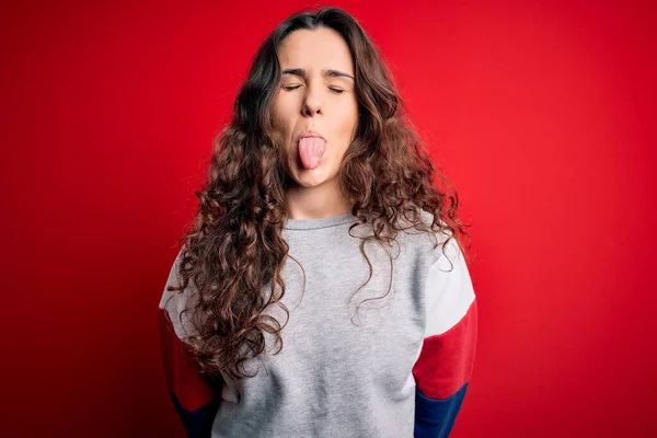 Young beautiful woman with curly hair wearing casual sweatshirt over isolated red background sticking tongue out happy with funny expression. Emotion concept.