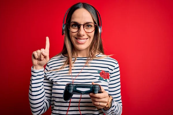 Young blonde gamer woman using gamepad playing video games over red background surprised with an idea or question pointing finger with happy face, number one