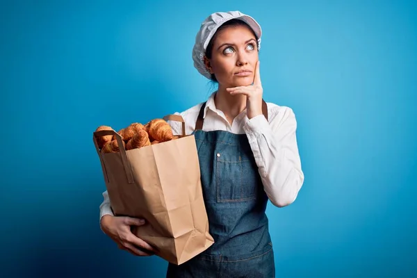 年轻美丽的面包师女人 蓝眼睛 头戴围裙 拿着纸袋 脸上挂着羊角面包 严肃地思考着问题 非常迷惑的想法 — 图库照片