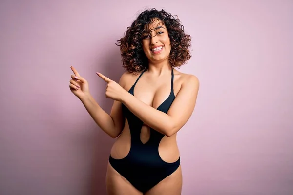 年轻美丽的阿拉伯女人穿着泳衣 戴着墨镜 带着粉红的背景 微笑着 看着摄像机 用双手和手指指向旁边 — 图库照片