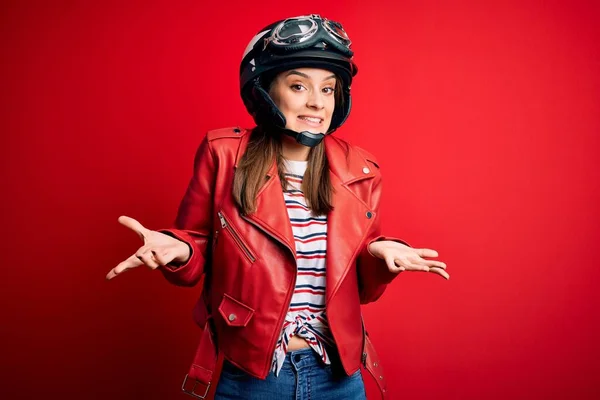 オートバイのヘルメットと赤いジャケットを身に着けている若い美しいブルネットの元サイクリストの女性は腕と手を上げて混乱した表情 疑いの概念 — ストック写真