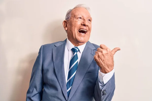 Senior handsome grey-haired businessman wearing elegant suit over white background pointing thumb up to the side smiling happy with open mouth