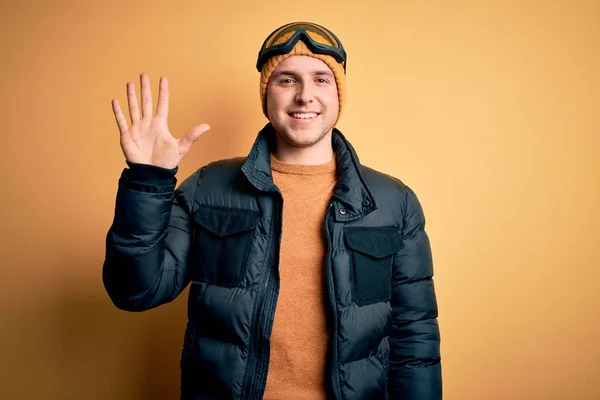 年轻英俊的高加索男子 戴着帽子 外套和滑雪眼镜 在冬雪天气里 他带着五号手指 面带微笑 自信而快乐 — 图库照片