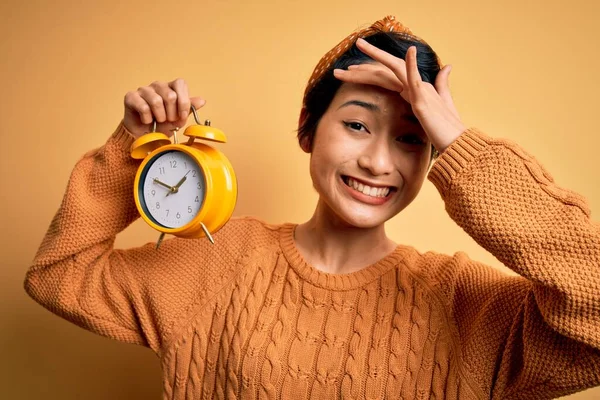 Young beautiful chinese woman holding vintage alarm clock over isolated yellow background stressed with hand on head, shocked with shame and surprise face, angry and frustrated. Fear and upset for mistake.
