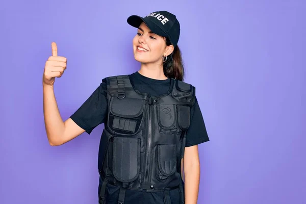 Young police woman wearing security bulletproof vest uniform over purple background Looking proud, smiling doing thumbs up gesture to the side