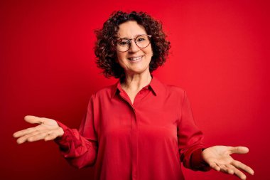 Orta yaşlı güzel kıvırcık saçlı kadın kırmızı arka planda günlük gömlek ve gözlük takıyor. Neşeli gülümsüyor ve kollarını açıyor. Dostça karşılama, olumlu ve kendinden emin selamlar.