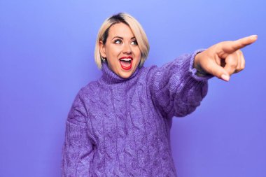 Güzel sarışın ve cüsseli bir kadın mor arkaplan üzerine balıkçı yaka kazak giyiyor. Parmağı önde, ağzı açık, şaşırmış bir ifade sergiliyor.