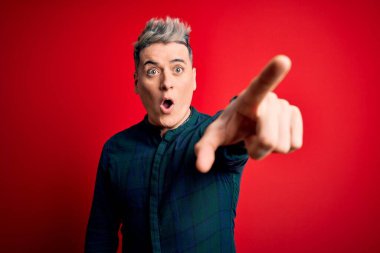 Kırmızı izole edilmiş arka plan üzerinde zarif yeşil tişört giyen genç, yakışıklı, modern bir adam. Parmakları önde, ağzı açık, şaşırmış, ön tarafta bir şey var.