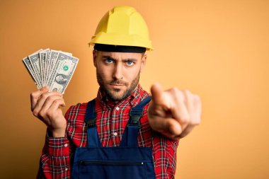 Güvenlik kaskı takan genç müteahhit sarı zemin üzerinde dolar tutarak kamerayı işaret ediyor ve sen, el işareti, ön taraftan pozitif ve kendinden emin bir jest.