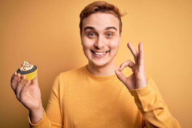 Genç ve yakışıklı kızıl saçlı adam sarı zemin üzerinde tatlı çikolatalı kek yiyor. Parmaklarıyla işaret veriyor, gülümseyerek mükemmel bir sembol oluşturuyor.