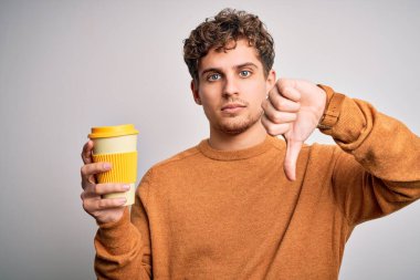 Sarışın genç adam, kıvırcık saçlı, beyaz arka planda duran kızgın yüzlü bir fincan kahve içiyor. Negatif işareti, baş parmakları aşağıda, reddedilme kavramı.
