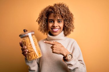 Genç Afro-Amerikan kıvırcık kadın elinde kavanozla İtalyan makarnası tutarken mutlu mesut elini ve parmağını gösterdi.