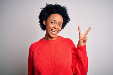 Kıvırcık saçlı, kırmızı süveter giyen, kameraya gülümseyen ve parmaklarıyla zafer işareti yapan güzel Afro-Amerikan kadın. İki numara..