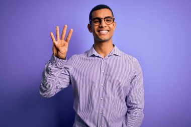 Yakışıklı Afro-Amerikalı adam mor arka planda çizgili gömlek ve gözlük takıyor ve parmaklarını dört numarayı gösterirken kendinden emin ve mutlu gülümsüyor..