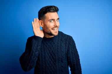 İzole edilmiş mavi arka planda dikilen sıradan bir süveter giyen genç yakışıklı bir adam. Kulağı kulağında, dedikoduları ve dedikoduları dinliyor. Sağırlık kavramı.