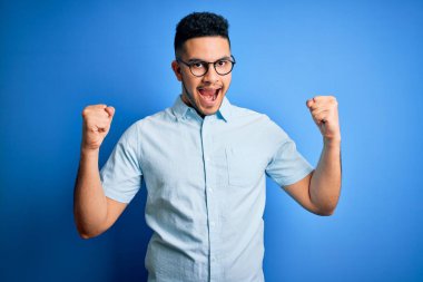 Genç ve yakışıklı bir adam günlük gömleği ve gözlükleriyle mavi arka plan üzerinde şaşırmış ve şaşırmış bir şekilde kollarını kaldırarak ve gözlerini açarak başarıyı kutluyor. Kazanan konsept.