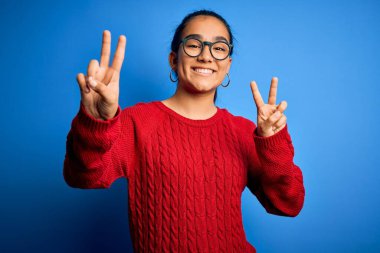 Genç, güzel Asyalı bir kadın mavi arka planda gözlük ve süveter takmış kameraya bakıp zafer işareti yaparken gülümsüyor. İki numara..
