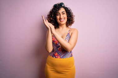 Genç, güzel bir Arap kadın, mayo ve pembe arka planda güneş gözlüğü takıp mutlu ve neşeli alkışlıyor, gururla gülümsüyor.