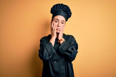 Güzel aşçı üniforması ve şapka giyen genç bir kadın sarı arka planda duruyor. Yüzü, depresyonu ve üzüntüyü örten yorgun eller, sorun yüzünden üzgün ve sinirli.