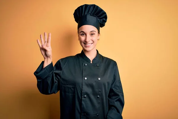 年轻美丽的厨师女士身穿炊事服 头戴一顶帽子 站在黄色的背景上 带着自信和快乐的笑容 用手指着四号 — 图库照片
