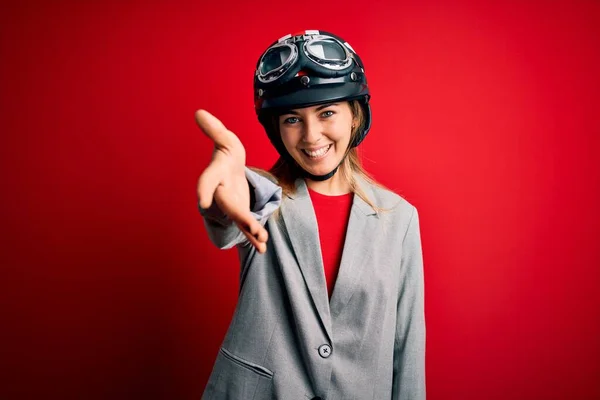 赤の背景にオートバイのヘルメットを身に着けている若い美しいブロンドのオートバイの女性は 挨拶や歓迎として握手を提供フレンドリーな笑みを浮かべて 成功事業 — ストック写真