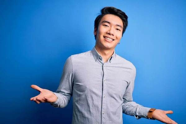 Young handsome chinese man wearing casual shirt standing over isolated blue background smiling cheerful with open arms as friendly welcome, positive and confident greetings