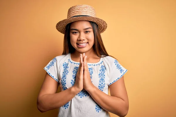 年轻美丽的亚洲女孩 身穿休闲装 头戴一顶帽子 站在黄色的背景上 手牵手祈祷 祈求宽恕 充满自信地微笑着 — 图库照片