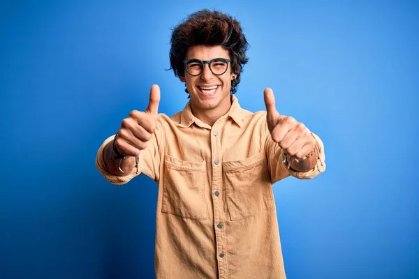 孤立した青い背景の上に立つカジュアルなシャツを着た若いハンサムな男は 手で積極的なジェスチャーを行うことを承認し 親指を上げて笑顔と成功のために幸せ 勝者のジェスチャー — ストック写真