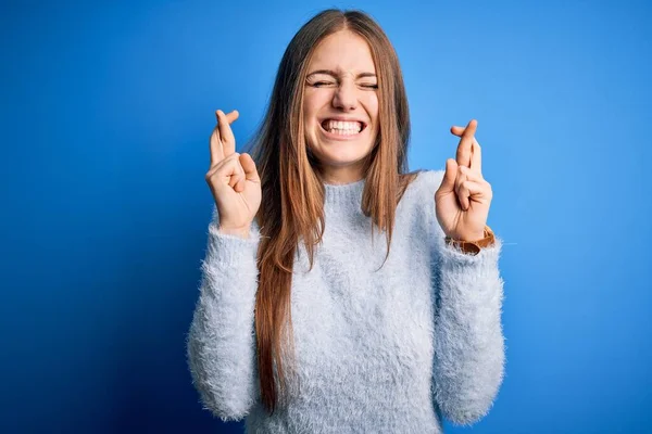 年轻美丽的红头发女人穿着休闲衫 戴在孤立的蓝色背景上 摆出手势 微笑着 合上了希望和眼睛 运气和迷信观念 — 图库照片