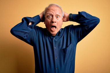 Gri saçlı, mavi gömlekli, sarı arka planda dikilen, elleri kafasında, korkmuş, ağzı açık, şok geçirmekten korkan ve şaşırmış yaşlı bir adam.