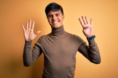 Sarı arka plan üzerine balıkçı yaka kazak giyen genç yakışıklı bir adam dokuz numaralı parmaklarını gösterirken kendinden emin ve mutlu bir şekilde gülümsüyor..