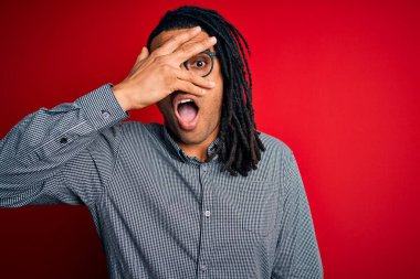 Young handsome african american man with dreadlocks wearing casual shirt and glasses peeking in shock covering face and eyes with hand, looking through fingers with embarrassed expression.