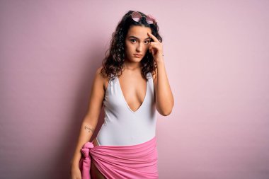 Tatilde kıvırcık saçlı, pembe arka planda beyaz mayo giyen güzel bir kadın alnındaki sivilceden mutsuz olduğunu gösteriyor. Siyah noktalı çirkin bir enfeksiyon. Sivilce ve deri problemi