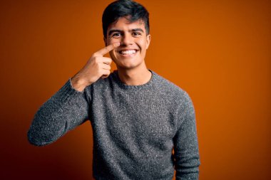 İzole edilmiş turuncu arka plan üzerinde duran, el parmağı ve burunla işaret eden, neşeli gülümseyen, gündelik süveter giyen genç yakışıklı bir adam. Güzellik konsepti