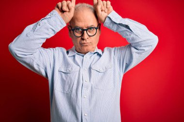 Orta yaşlı, yakışıklı, beyaz, sıradan çizgili tişört giyen ve kırmızı arka planda gözlük takan bir adam, boynuz gibi parmağıyla komik hareketler yapıyor.