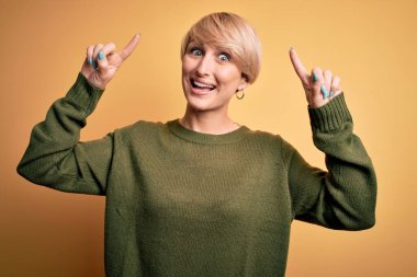 Modern kısa saçlı, sarı arka planda rahat süveter giyen genç sarışın kadın şaşırmış, şaşırmış ve parmaklarıyla işaret ediyor ve kollarını kaldırıyordu..