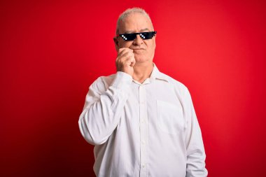 İzole edilmiş kırmızı arka planda komik güneş gözlüğü takan orta yaşlı, beyaz bir adam. Elleri çenesinde soru sormayı düşünüyor. Düşünceli bir ifade. Düşünceli bir yüzle gülümsüyor. Şüphe kavramı.