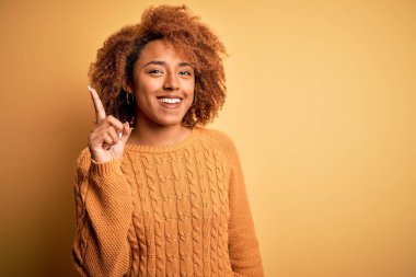 Kıvırcık saçlı, genç, Afro-Amerikan kadın. Günlük süveter giyiyor. Bir numarayı işaret ediyor ve gülümsüyor..