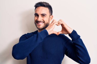 Sakallı, sıradan bir süveter giyen, beyaz arka planda duran, aşık bir genç adam elleriyle kalp sembolü çiziyor. Romantik konsept.