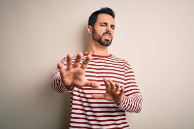 Sakallı, sıradan çizgili tişört giyen, beyaz arka planda duran, tiksindirici ve tiksindirici bakışları olan genç, yakışıklı bir adam. Eller havada