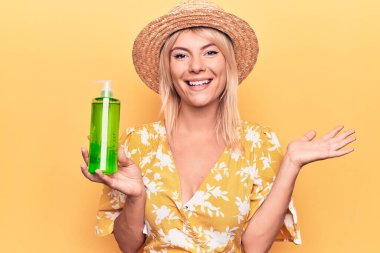 Yazlık şapkalı güzel sarışın kadın aloe vera kremalı bir şişe tutuyor başarıyı kutluyor mutlu bir gülümsemeyle ve kazanan yüz ifadesini kaldırarak.