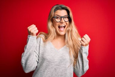 Genç, sarışın, süveter ve gözlük takan güzel bir kadın izole edilmiş kırmızı arka planda şaşırmış ve şaşırmış bir şekilde kollarını kaldırıp, gözlerini açarak başarıyı kutluyor. Kazanan konsept.
