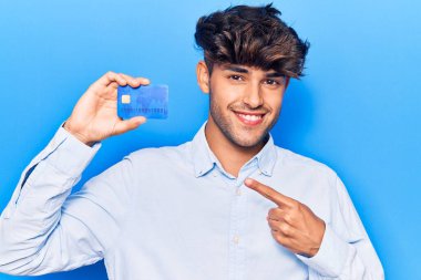 Kredi kartı tutan genç İspanyol adam mutlu bir şekilde el ve parmağıyla işaret ediyor. 