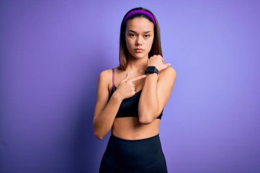 İzole edilmiş mor arka plan üzerine spor giyerek spor yapan genç ve güzel bir kız. Aceleyle saati, sabırsızlığı ve rahatlamış bir ifadeyle kameraya bakıyor.