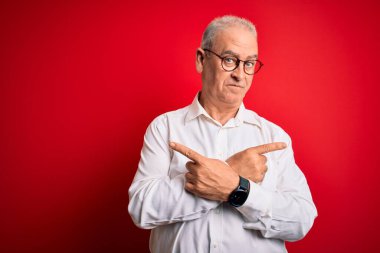 Orta yaşlı, beyaz gömlekli, kırmızı arka planda gözlüklü, yakışıklı bir adam. Her iki tarafı da işaret ediyor. Farklı yönler farklı.