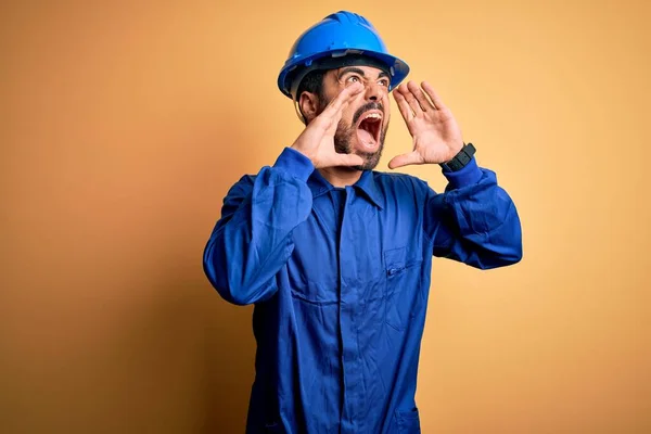 Mechanic man with beard wearing blue uniform and safety helmet over yellow background Shouting angry out loud with hands over mouth